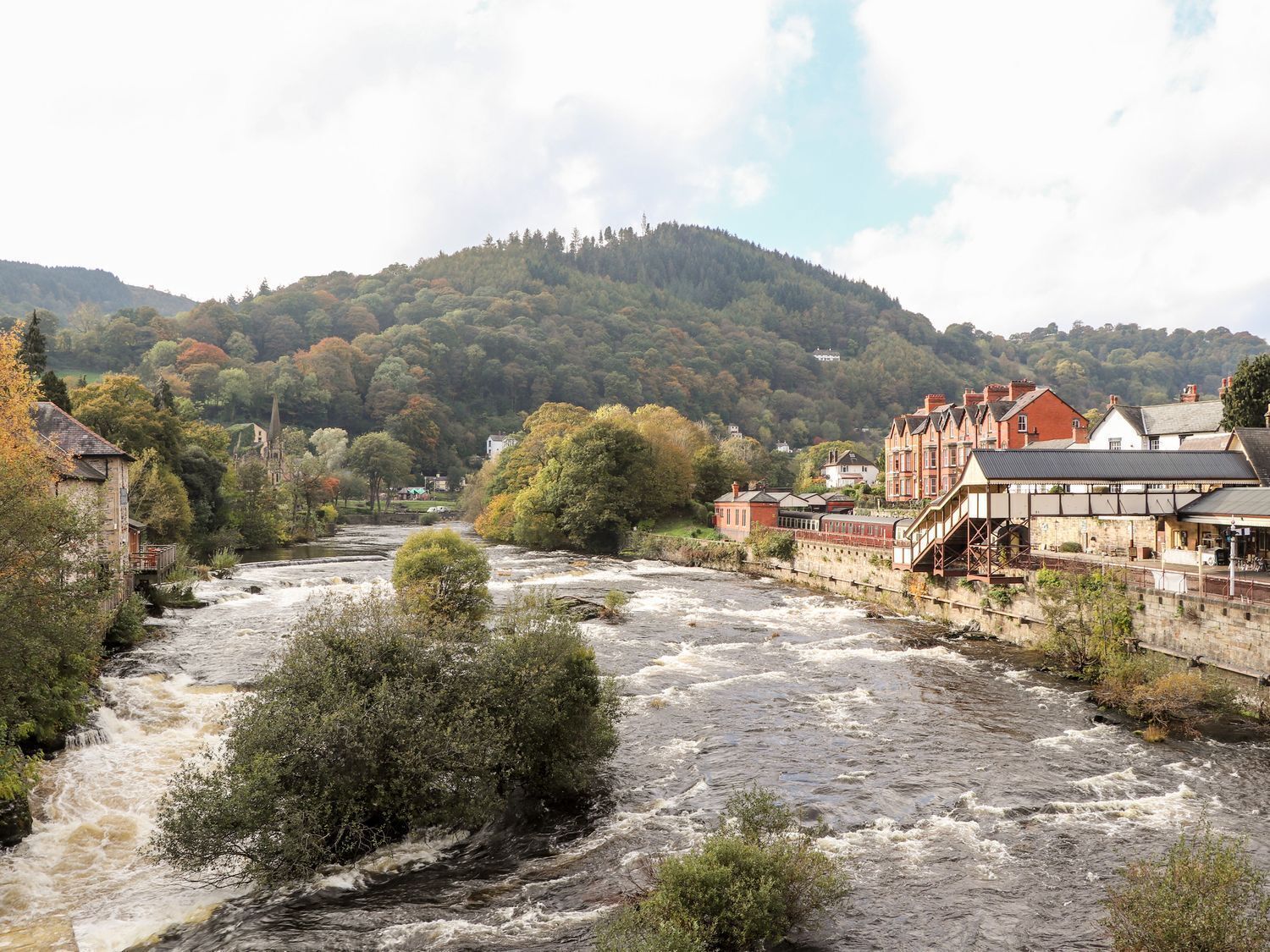 A river flowing through a town with buildings on the banks, a bridge, and a hill in the background.