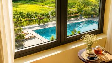 A room with a view of a swimming pool and lush greenery, a wooden chair with a cushion, and a small table with a vase and teapot.