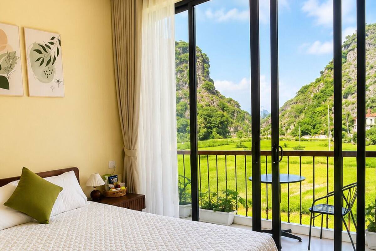 A bedroom with a large bed, a nightstand, a lamp, and a view of a balcony with a table and chairs.