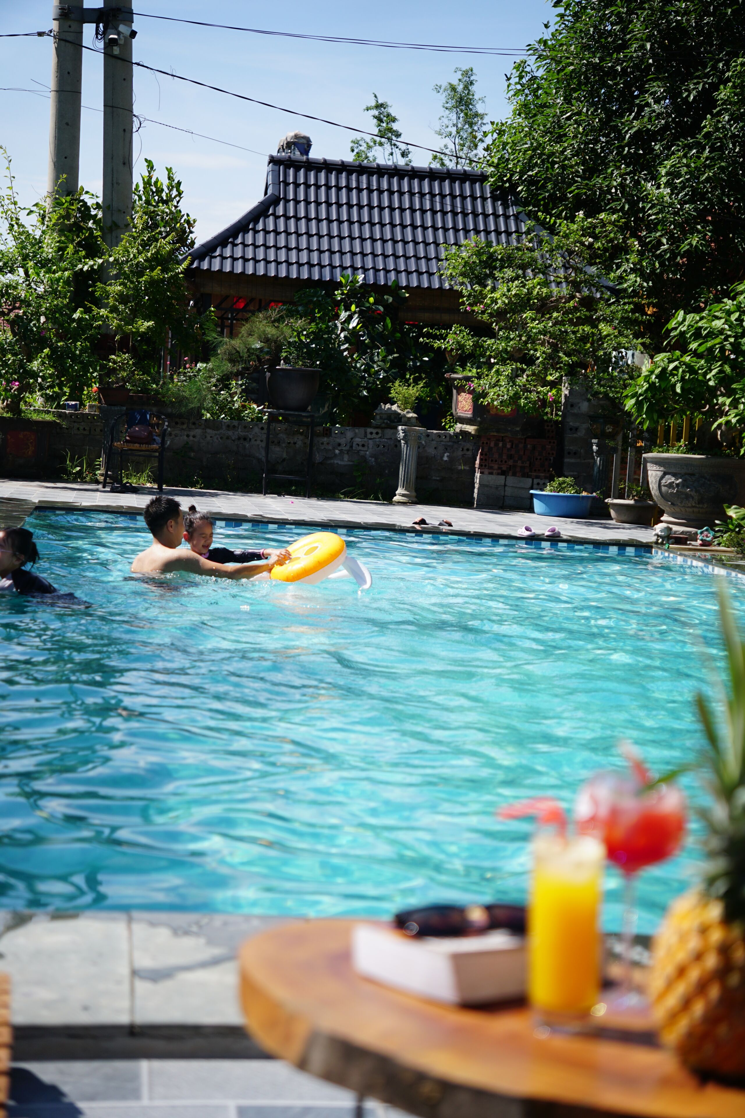 A swimming pool with people, a table with drinks, and a building with a tiled roof in the background.