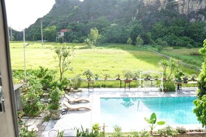 A swimming pool with a surrounding deck, a small garden area with potted plants, and a view of a green field and mountains in the background. - Tam Coc AppA House (Nam Hoa Lu)
