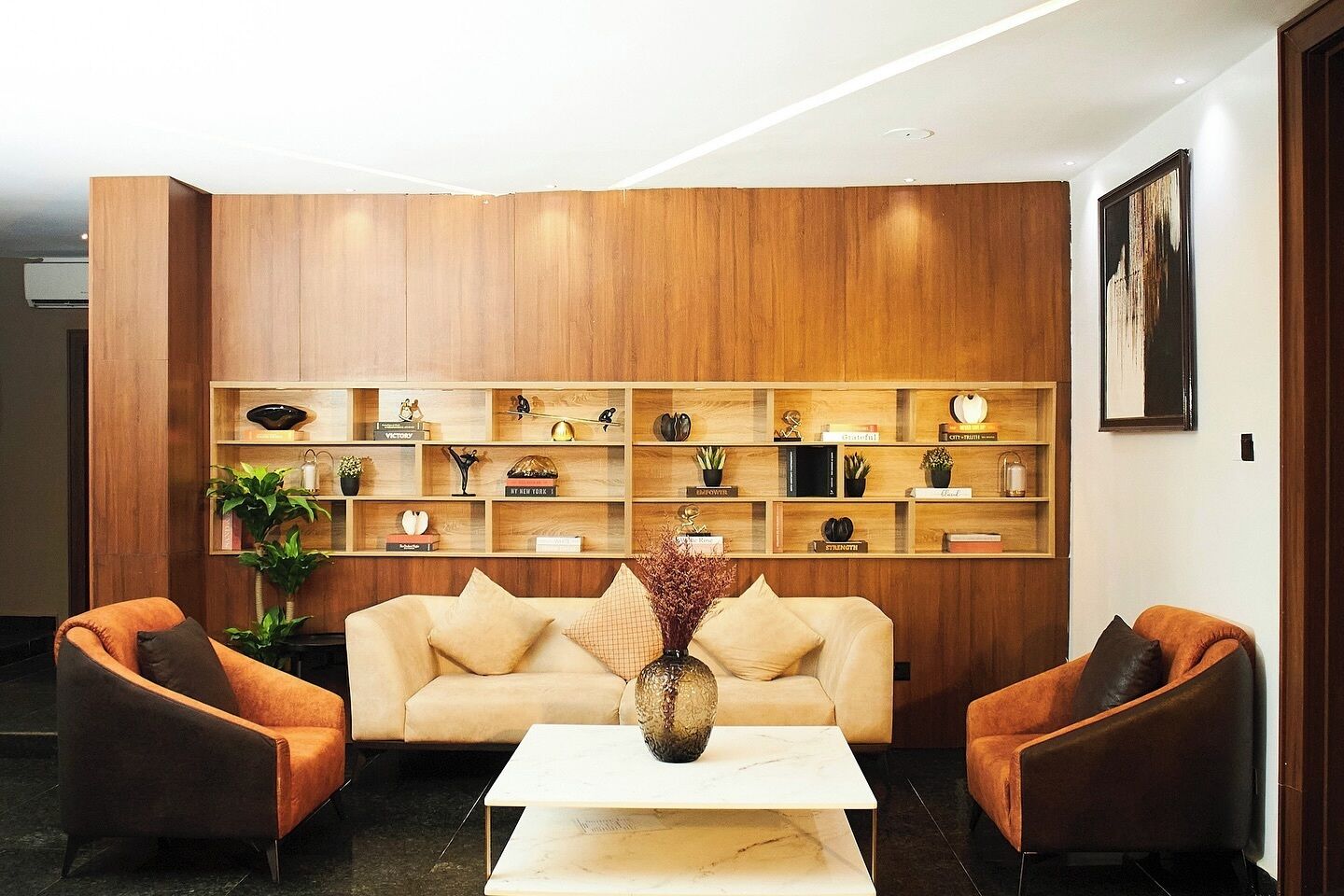 A modern living room with a wooden panel wall, a beige sofa, two armchairs, a marble coffee table, and a variety of decorative items on shelves.