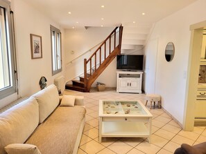 Un salon comprenant un canapé, une table basse agrémentée d’objets décoratifs, un téléviseur posé sur un meuble, un escalier et une vue sur l’extérieur.