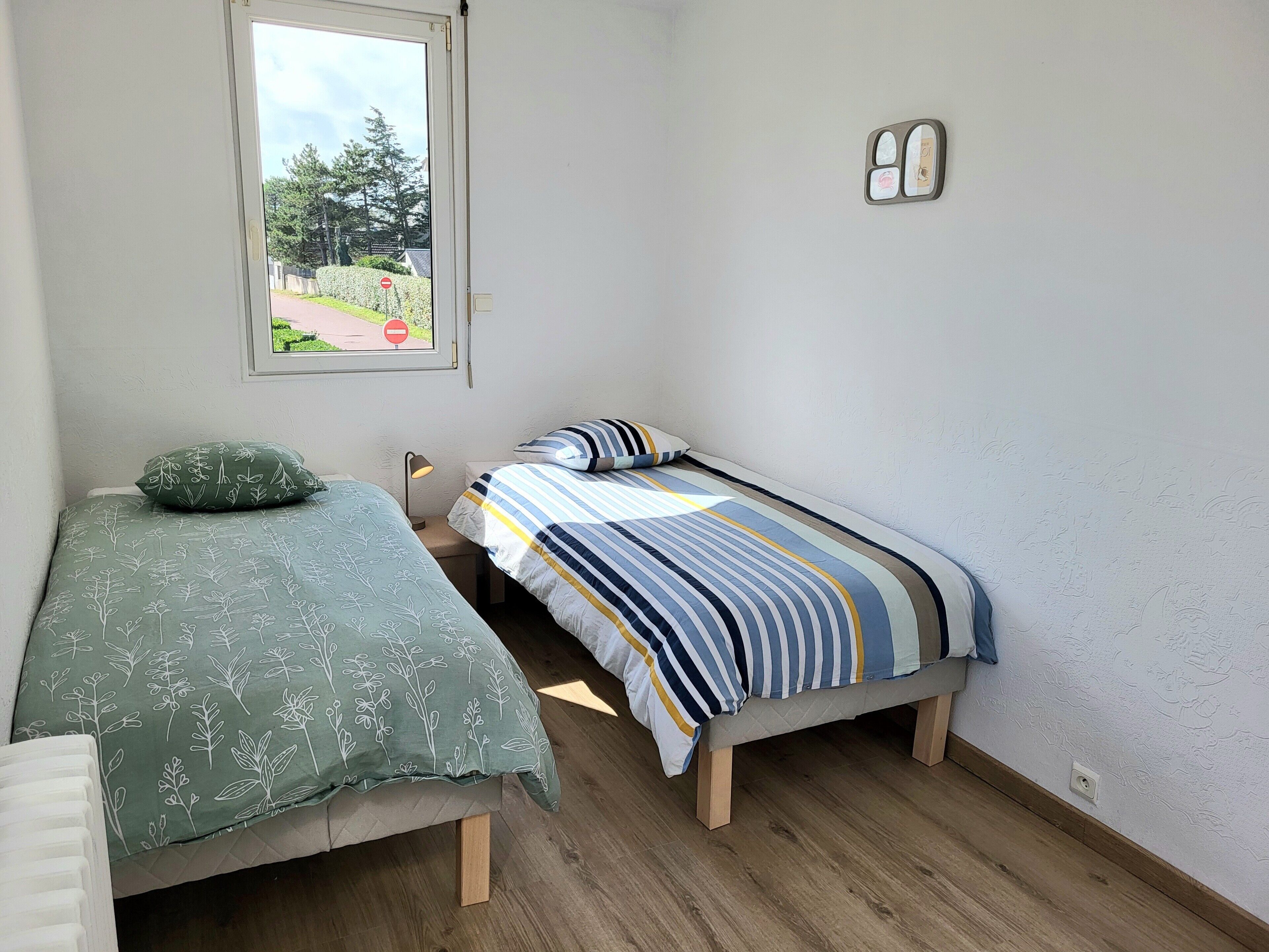 A small, clean room with two single beds, a window with a view of greenery, and a wall-mounted light fixture.