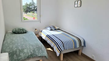 A small, clean room with two single beds, a window with a view of greenery, and a wall-mounted light fixture.