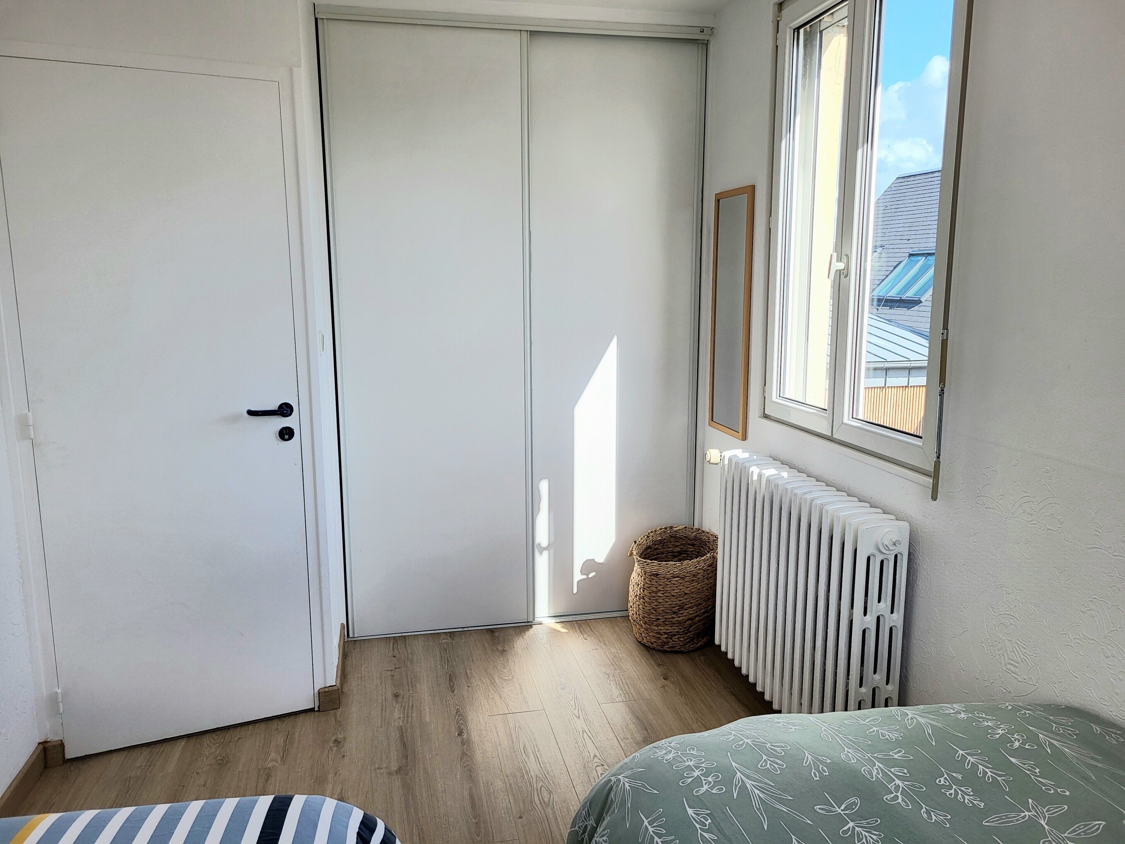 A room with a closed door, a radiator, a window with a view of a building, and a bed with a patterned bedspread.