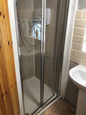 Una ducha con paredes de vidrio, un cabezal de ducha de mano, una puerta de madera y un lavamanos blanco con salpicadero de azulejos.
