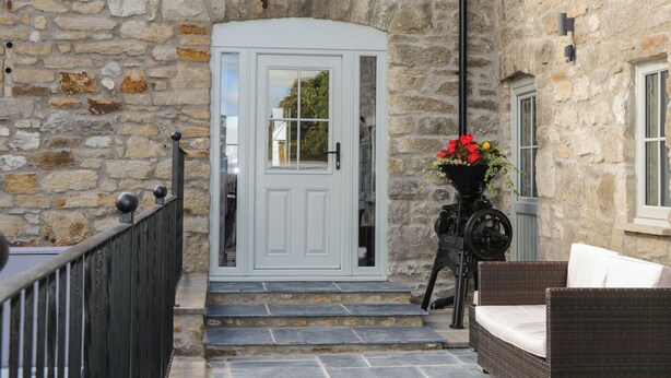 A stone building with a white door, steps leading to a porch, and a wicker chair. - Sun Trevor Barns 2 (Llangollen)