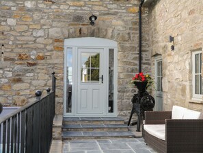 A stone building with a white door, steps leading to a porch, and a wicker chair. - Sun Trevor Barns 2 (Llangollen)