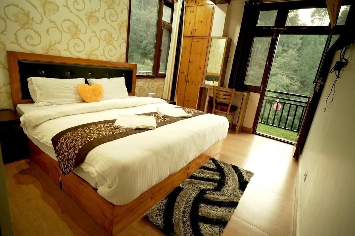 A bedroom with a large bed, a wooden headboard, a wardrobe, a desk, and a balcony with a view of greenery.