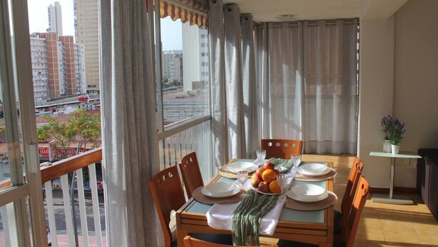 Un coin repas avec une table dressée pour deux personnes, une vue sur la ville et un balcon agrémenté de rideaux.