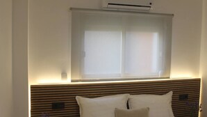 Une chambre d’hôtel avec un lit, une fenêtre avec des stores, un climatiseur fixé au mur et une tête de lit en bois.