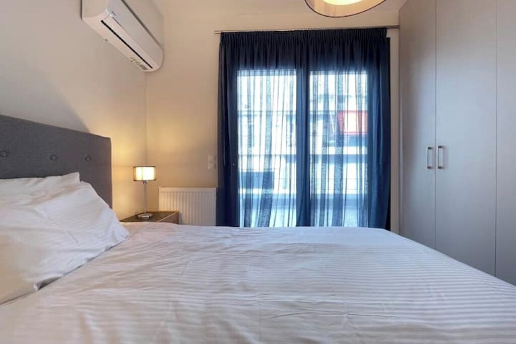 A hotel room with a bed, a nightstand, a lamp, an air conditioning unit, and a view through a window with curtains.
