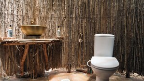 A rustic bathroom with a stone floor, a white toilet, a wooden basin, and a bamboo wall.