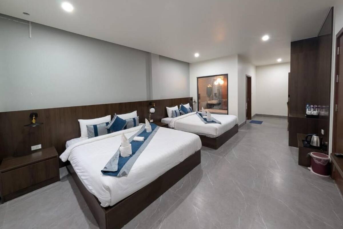 Une chambre d’hôtel avec deux lits, une tête de lit en bois, une table de chevet, un miroir, un tableau encadré, une petite table et un meuble contenant des articles de toilette.