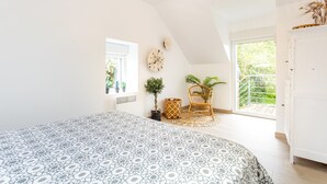 Une chambre à coucher avec un grand lit, une fenêtre, une plante, une chaise en osier et une armoire blanche.