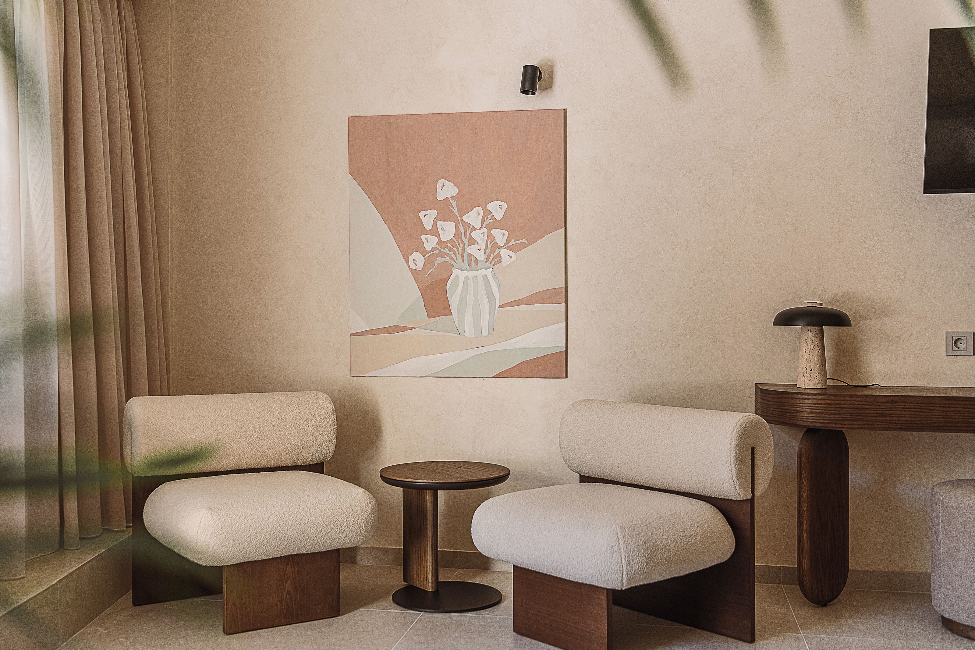 A room with two upholstered chairs, a small round table, a painting on the wall, and a lamp on a wooden side table.