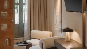 A modern hotel room with a sofa, a small table, a TV mounted on the wall, and a window with curtains. - Eteon Living Suites (Chania)