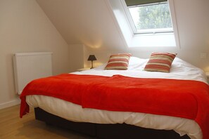 Quarto com teto inclinado, claraboia, radiador, abajur de cabeceira e cama com manta vermelha e almofadas listradas.