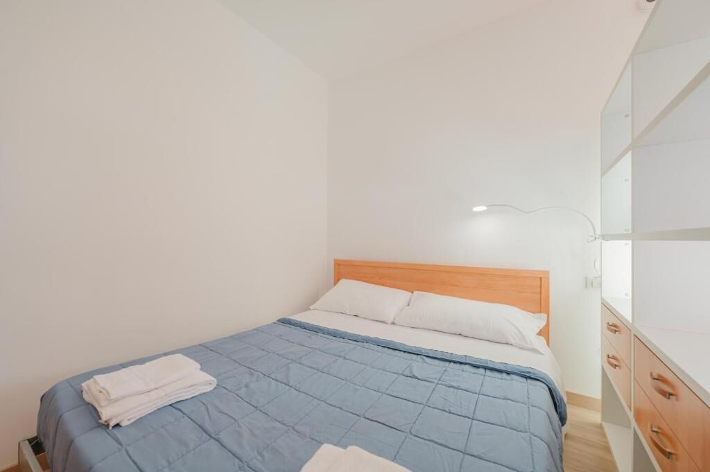Una cama individual con colcha azul, almohadas blancas y cabecera de madera. A la derecha, una pared blanca y un armario con cajones.