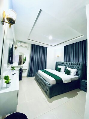 A modern bedroom with a tufted headboard bed, a white desk with a mirror, and a potted plant. - Bengerald Hotel And Suites (Abuja)