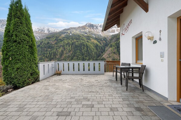 A balcony with a view of snow-capped mountains, a wooden table and chairs, and a white wall with a nameplate.