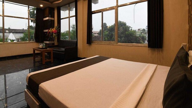 A hotel room with a large bed, a desk, a chair, a small table with a vase of flowers, and large windows offering a view of the outside. - Townhouse Aiims Kolkata Tanishq Inn (Kalyani)
