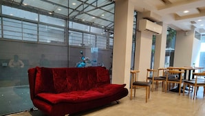 A red sofa, wooden chairs, and tables in a modern indoor space with large windows and an air conditioning unit. - Townhouse Aiims Kolkata Tanishq Inn (Kalyani)