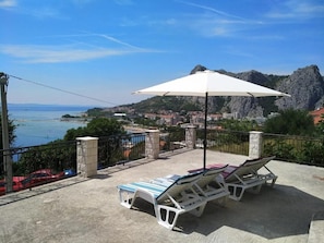 Ein Balkon mit Lounge-Möbeln und Sonnenschirm mit Blick auf eine Küstenstadt und Berge.