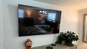 A flat-screen TV mounted on a wall with a logo and text indicating it's a music service. Below the TV, there's a decorative vase and a plant on a wooden cabinet. - Casa Blanca - Villa D11 (Sarasota)