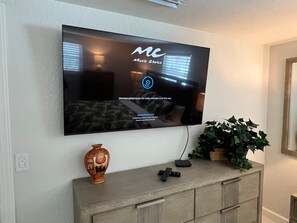 A flat-screen TV mounted on a wall with a logo and text indicating it's a music service. Below the TV, there's a decorative vase and a plant on a wooden cabinet. - Casa Blanca - Villa D11 (Sarasota)
