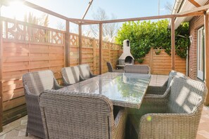 Une terrasse couverte avec une table à plateau de verre et des chaises en osier, un mur en briques et une clôture en bois.