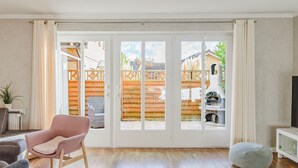 Ein geräumiger Raum mit einer Glasschiebetür, die zu einem Garten führt, ein rosa Sessel, ein grauer Hocker und ein weißes Schranksystem.