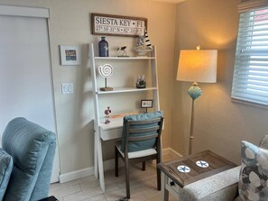 A cozy room with a white desk, a chair, a small table, and a lamp. There is a sign on the wall and a window with blinds. - Casa Blanca - Villa D03 (Sarasota)