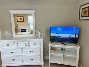 A white dresser with drawers and a mirror above it, a television on a stand with a beach scene, and a framed picture on the wall. - Casa Blanca  -Villa C15 (Sarasota)