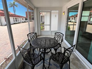 A covered patio with a round table and four chairs, a sliding glass door, and a view of a residential area with palm trees and houses. - Casa Blanca  -Villa C15 (Sarasota)