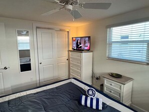 Ein Schlafzimmer mit einem Bett, einem Fernseher auf einer Kommode, einem Deckenventilator und einem Fenster mit Jalousien.