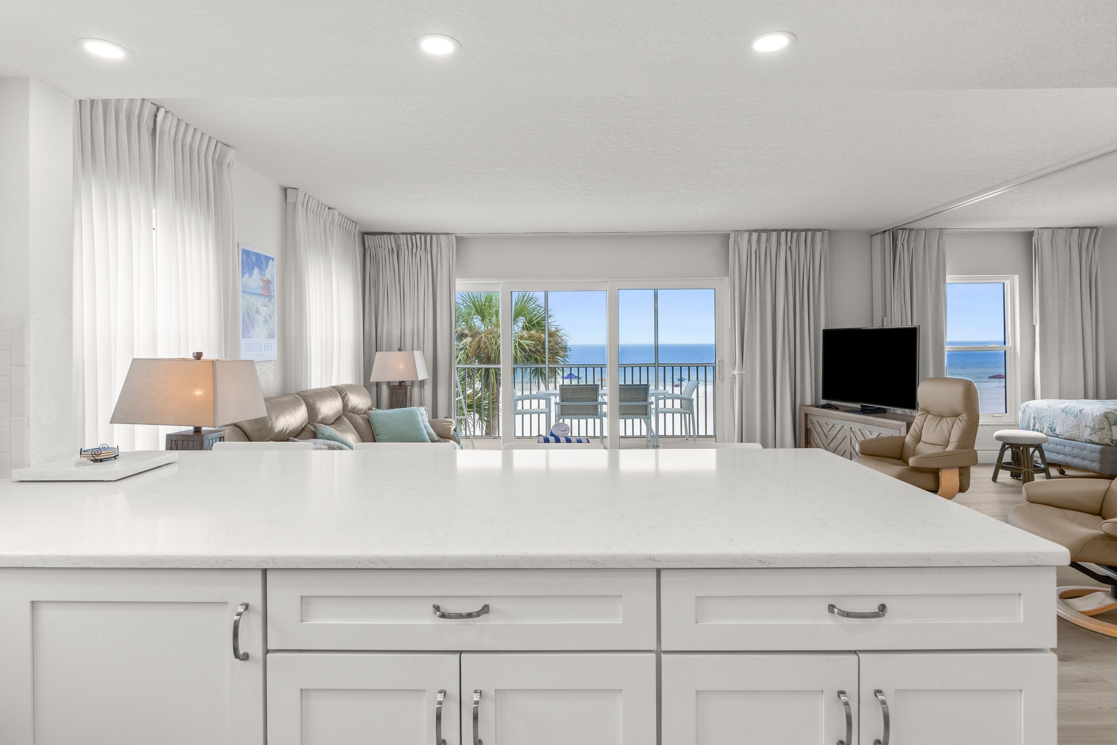 A spacious living area with a large white island, a flat-screen TV, and a view of the sea through the balcony doors.