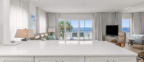 A spacious living area with a large white island, a flat-screen TV, and a view of the sea through the balcony doors.