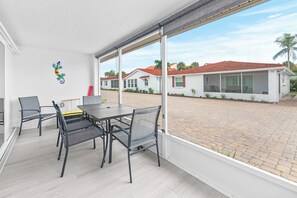 A covered outdoor area with a table and chairs, a view of a residential street, and a decorative wall piece. - Casa Blanca - Villa C06 (Sarasota)