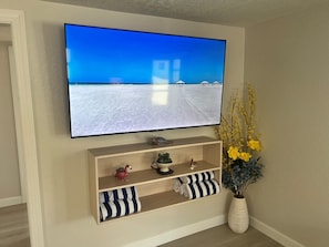 A television mounted on a wall with a beach scene, a wooden shelf with decorative items, and a vase with yellow flowers. - C06 - Where Island Dreams Come to Life (Sarasota)
