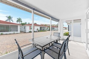 A covered outdoor patio with a table and chairs, a view of a residential area with houses and palm trees, and a door leading to the interior. - Casa Blanca - Villa C06 (Sarasota)