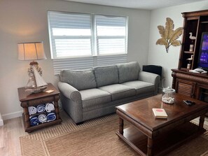 Ein Wohnzimmer mit grauem Sofa, einem Holztisch, einer Lampe, einer Zierpflanze und einem Fernseher.