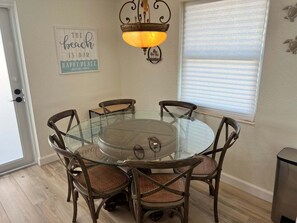 A dining area with a glass table surrounded by wooden chairs with woven seats, a wall-mounted light fixture, and a sign with a beach message. - Casa Blanca - Villa C05 (Sarasota)