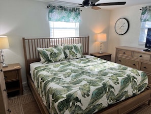 A bedroom with a bed, two bedside tables, a nightstand, a ceiling fan, a wall clock, and a window with curtains. - Casa Blanca - Villa C05 (Sarasota)