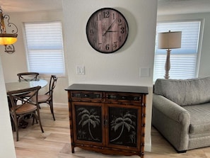 A living room with a round wall clock, a wooden cabinet with palm tree designs, a gray sofa, and a dining area with chairs and a table. - Casa Blanca - Villa C05 (Sarasota)