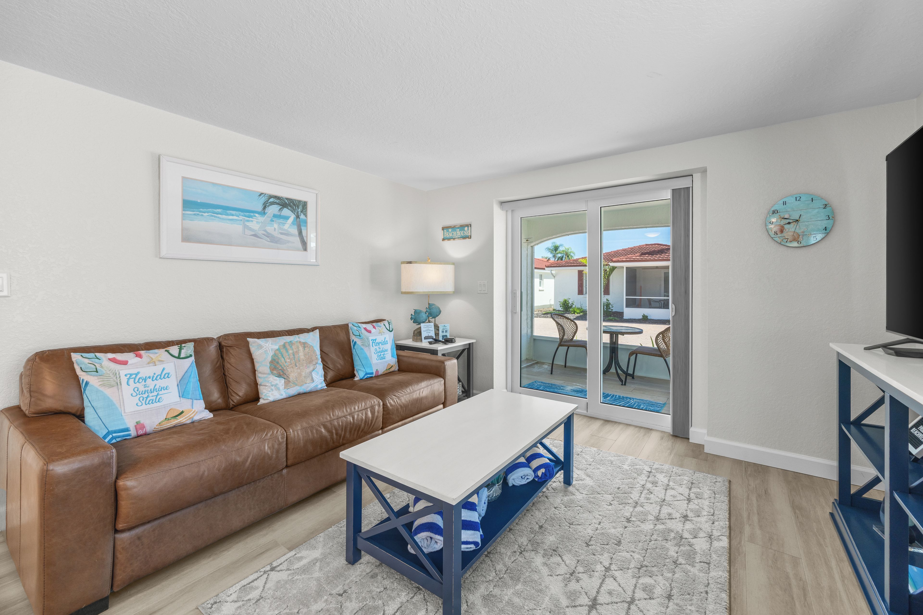 A living room with a brown leather sofa, a white coffee table, and a blue and white patterned rug. There is a painting of a beach scene on the wall, a lamp, and a clock.