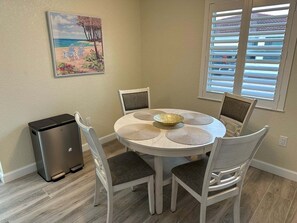 A dining area with a round table, four chairs, a small refrigerator, and a window with blinds. - Casa Blanca - Villa A12 (Sarasota)