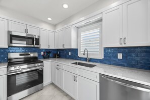A modern kitchen with white cabinets, stainless steel appliances, a blue backsplash, and a window with blinds. - Casa Blanca A18- Siesta Key Is Calling (Sarasota)