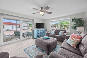 A spacious living room with a ceiling fan, a flat-screen TV, and a large window offering a view of a residential area. - Casa Blanca - Villa B01 (Sarasota)